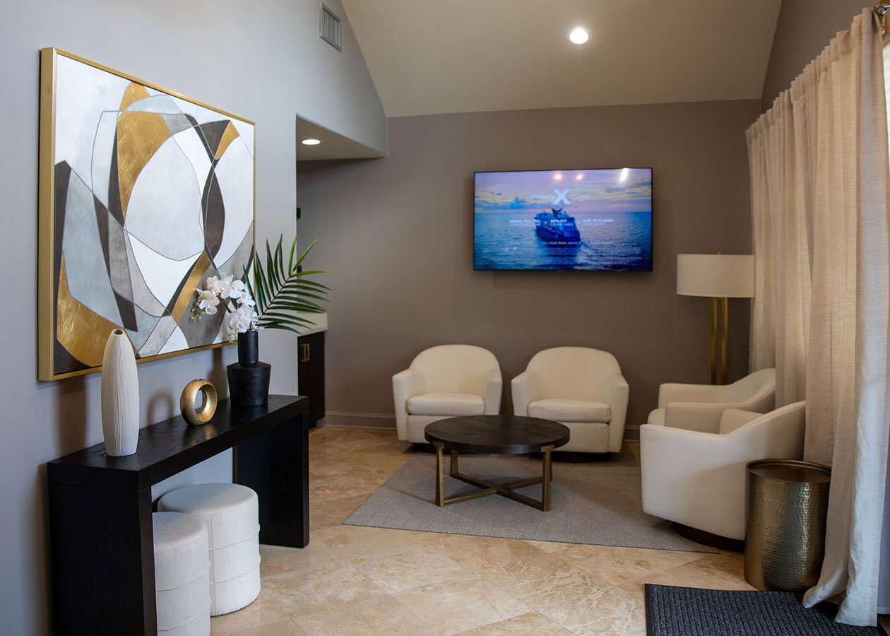 A modern living room with beige tile flooring, white armchairs, a round dark wood coffee table on a gray rug, a wall-mounted television displaying a cruise ship, a tall beige curtain, a gold and black floor lamp, a black console table with decorative