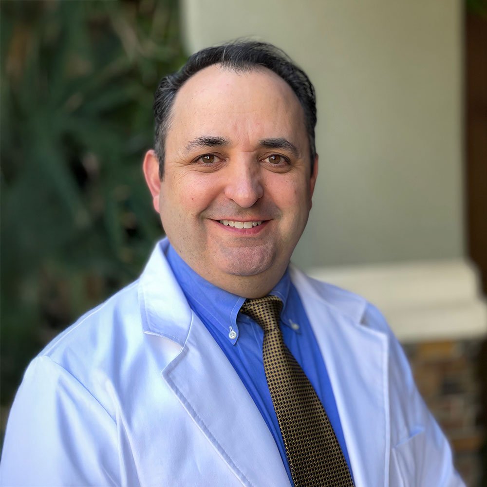 A smiling man in a white coat, blue dress shirt, and patterned tie outdoors with greenery in the background.