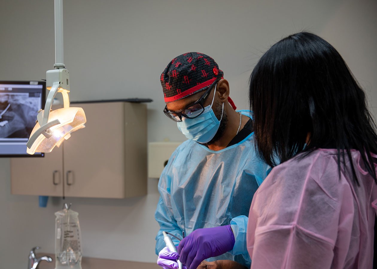 Dentists in protective gear performing a dental procedure in a clinic with a monitor displaying an X-ray and various dental tools.